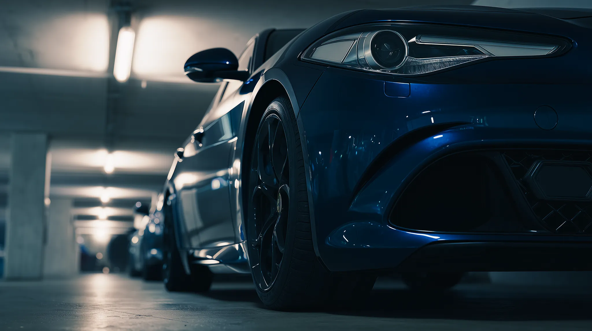 Vista ravvicinata della ruota anteriore e del faro di un'auto sportiva blu in un parcheggio sotterraneo illuminato.