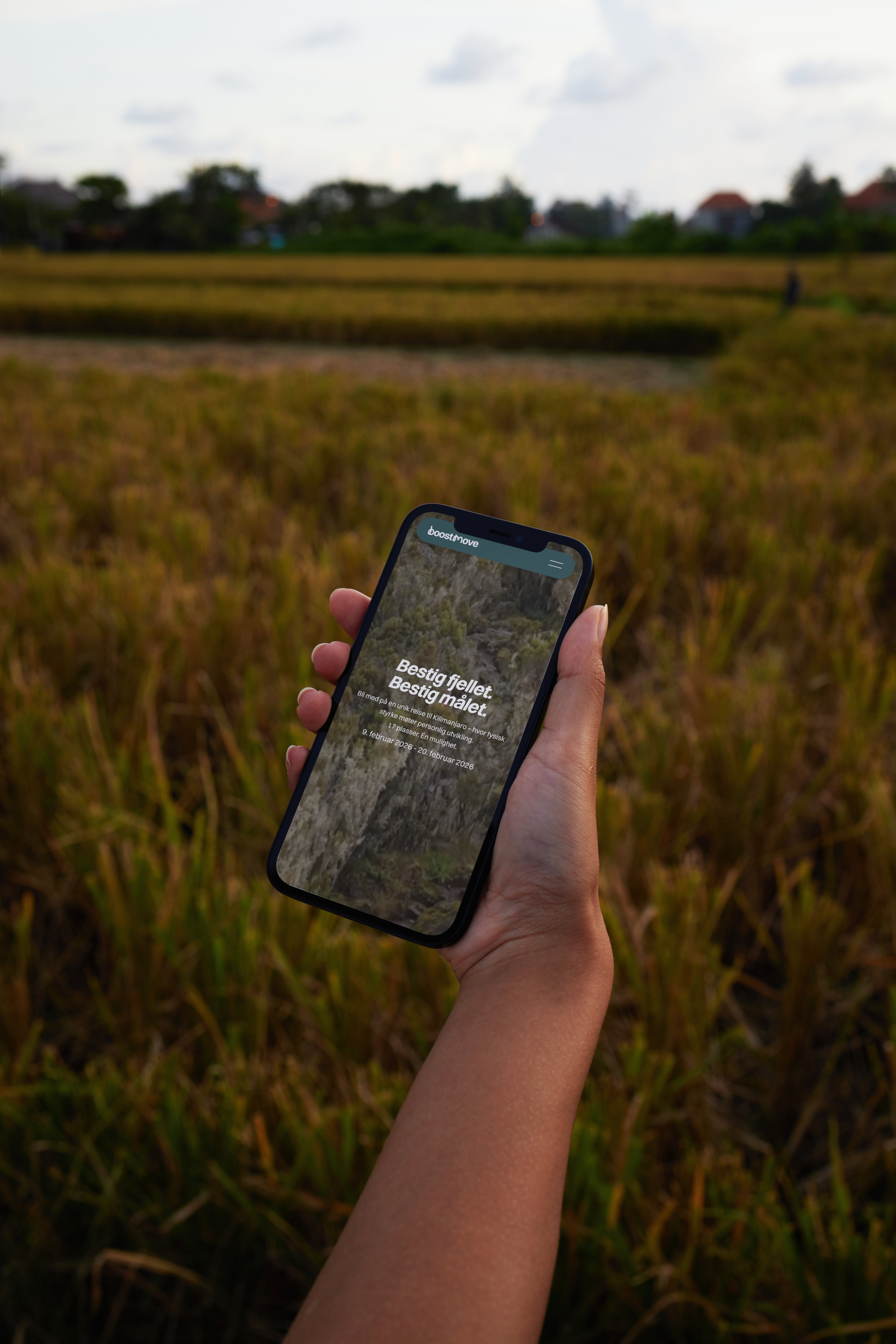 Hånd som holder en mobiltelefon som viser en nettside med teksten 'Bestig fjellet. Bestig målet.' i et utendørs landskap med gress og trær.