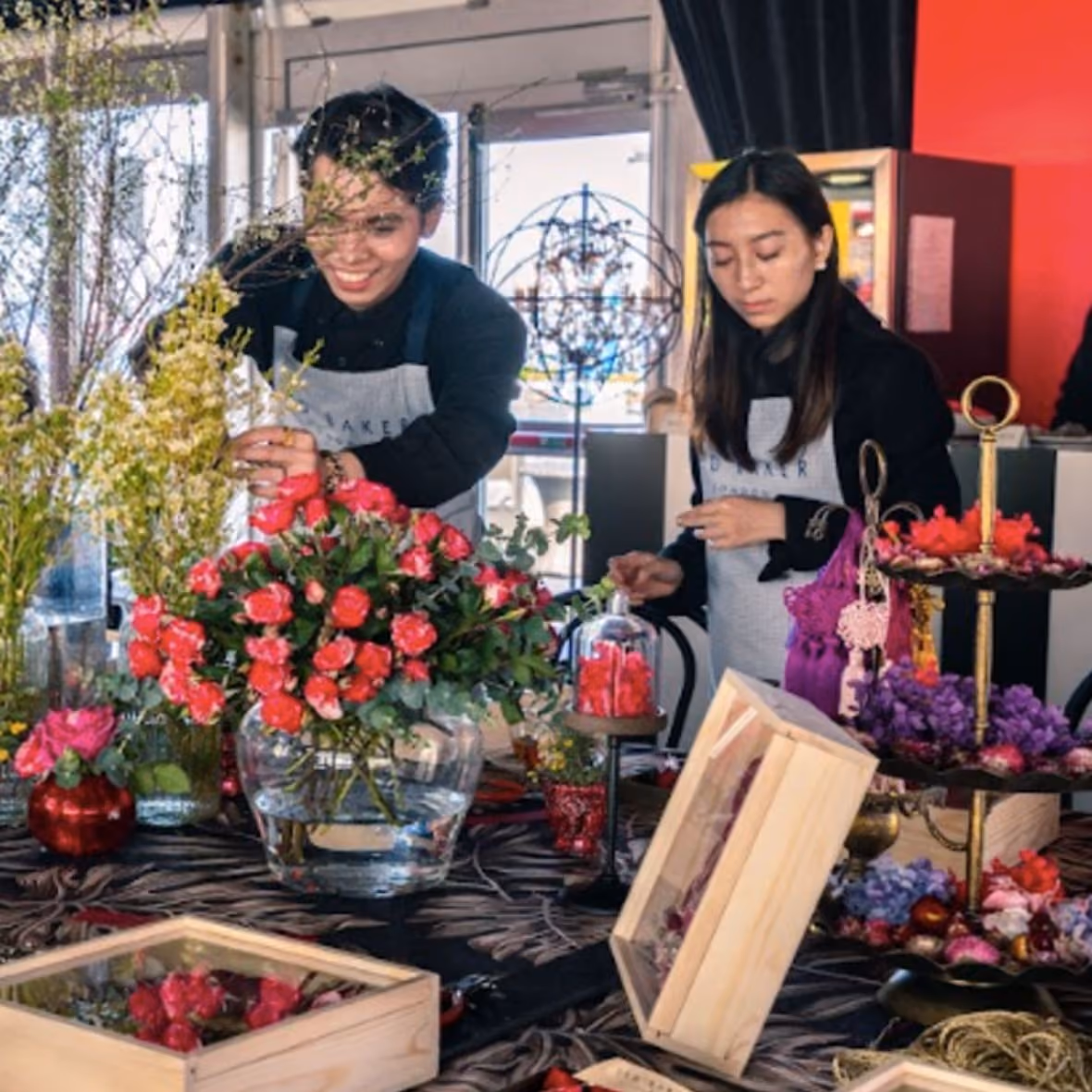 Two people arranging colorful flowers on a table filled with floral decorations and vases inside a room.