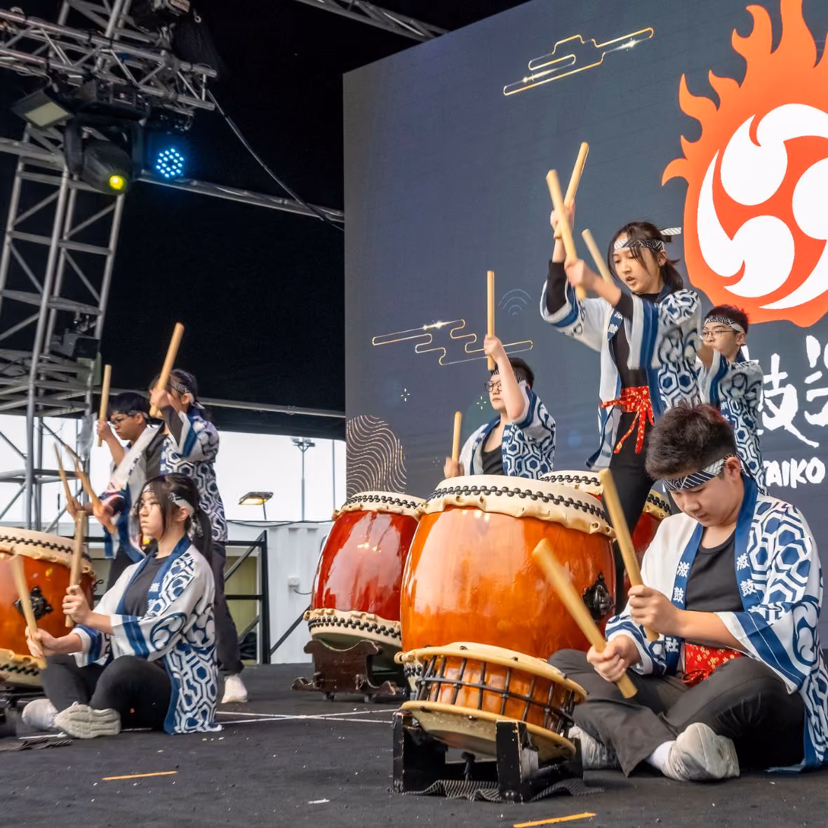 A group of young performers playing traditional Japanese taiko drums on stage, dressed in patterned blue and white festival clothing with headbands.
