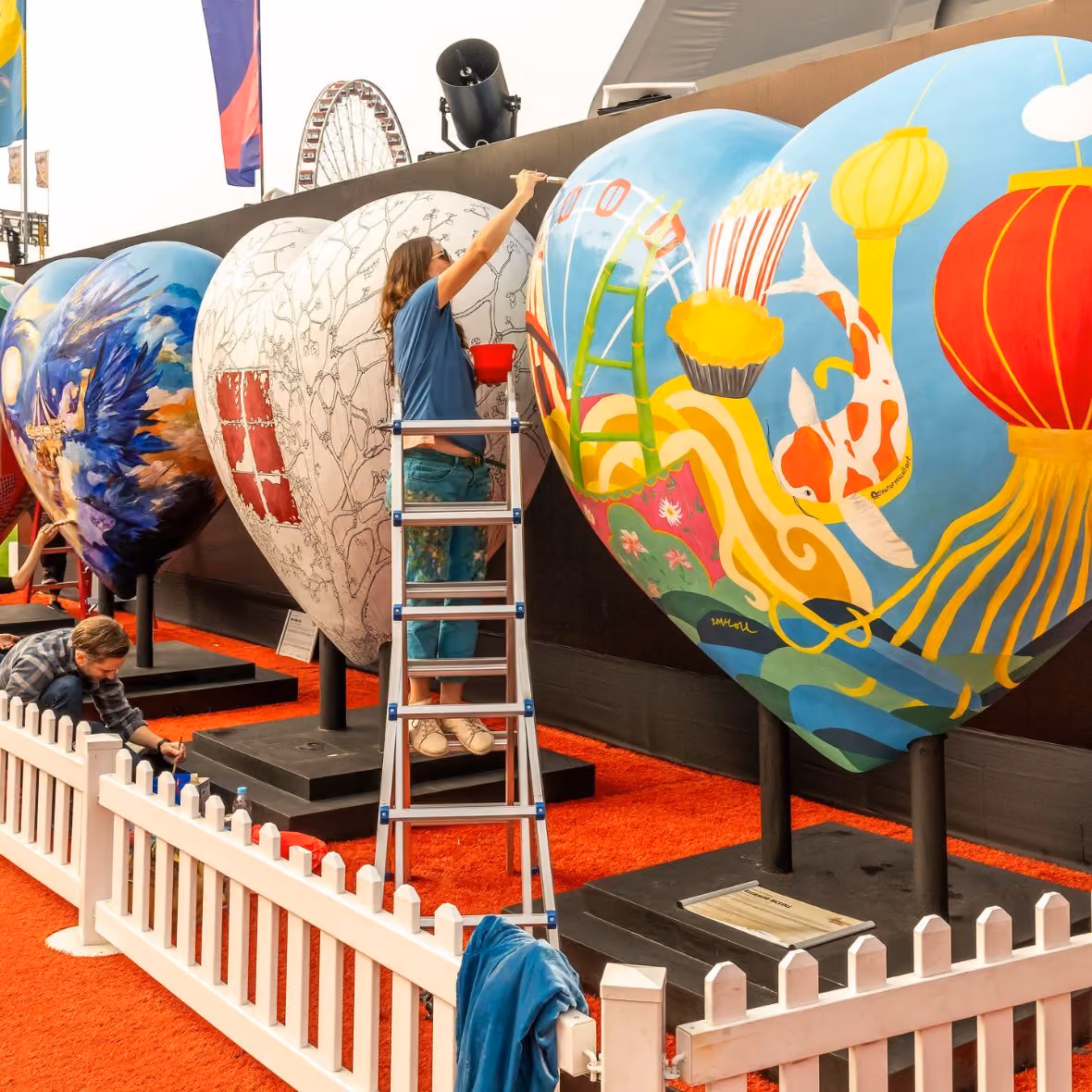 Two artists painting large heart-shaped sculptures with colorful designs at an outdoor event.