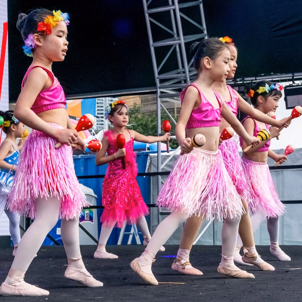Young girls in pink tops, pink skirts, and floral headbands performing a dance with maracas on stage.