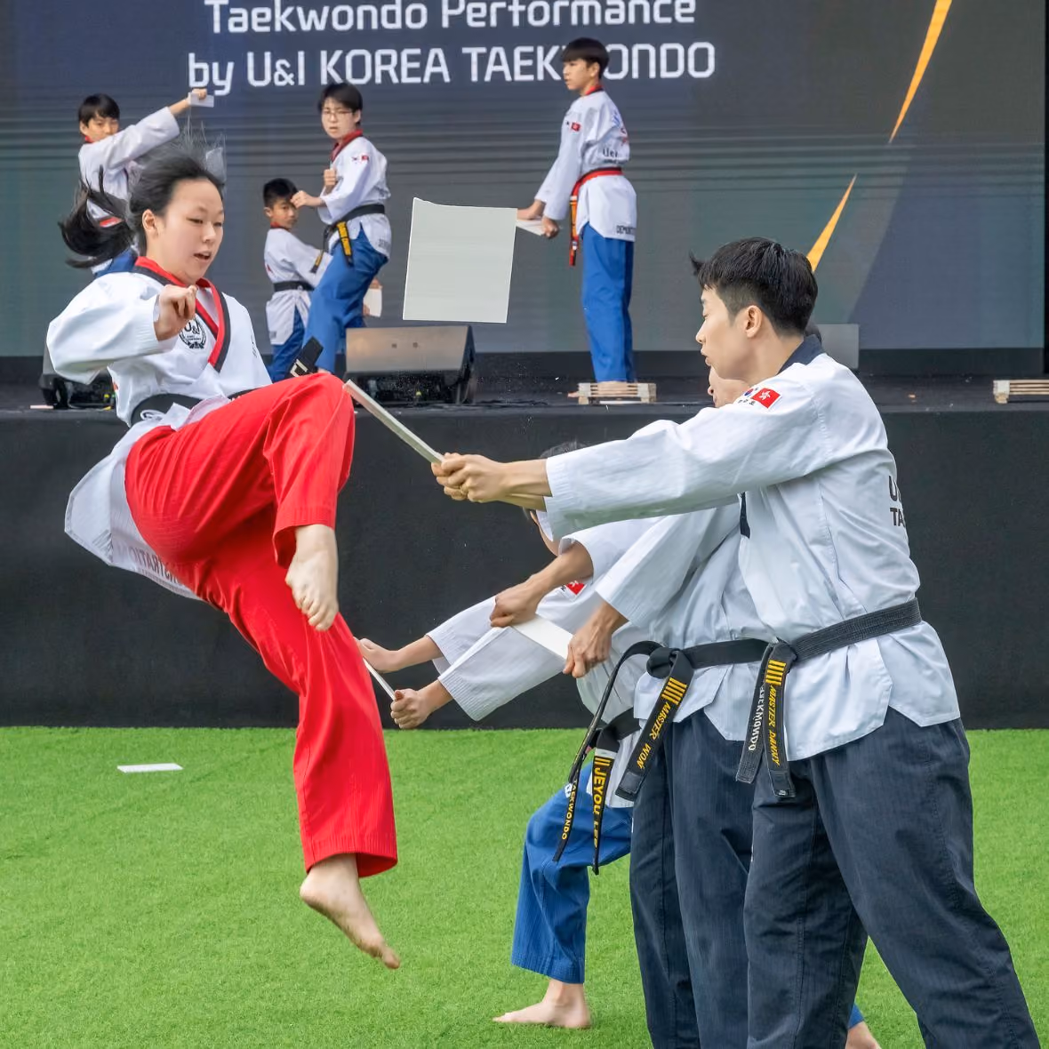 Female taekwondo martial artist in red pants breaking a board held by two black belt practitioners during a performance.