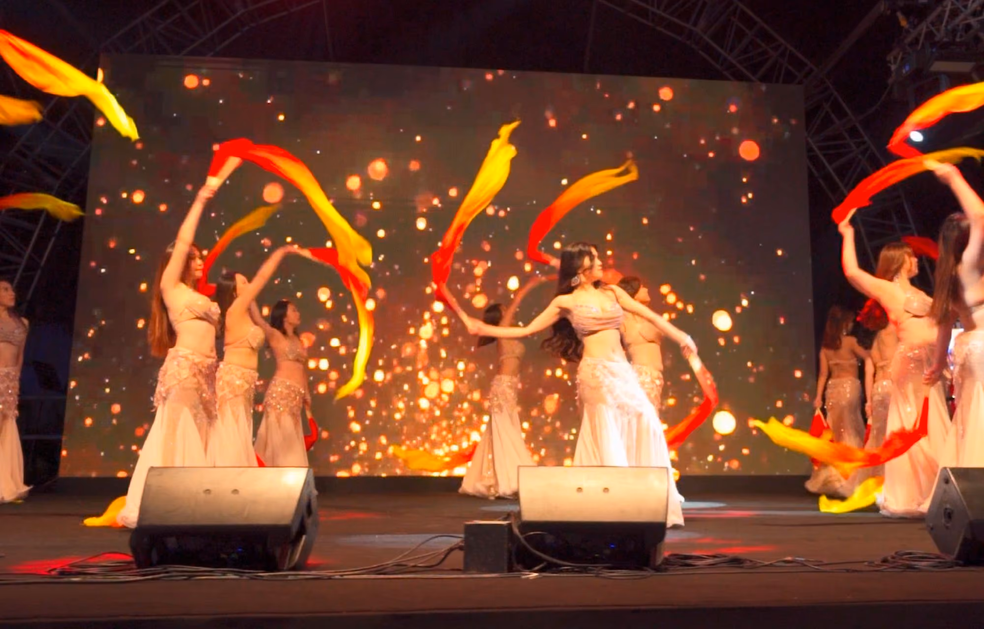 Group of women performing a choreographed dance on stage with flowing red and yellow scarves against a backdrop of sparkling lights.