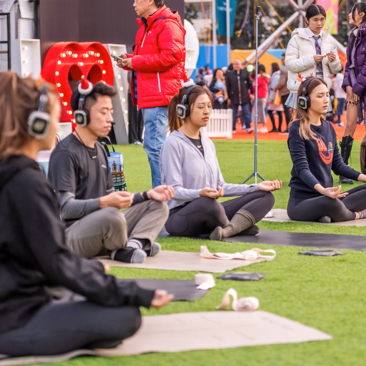 Group of young people meditating outdoors on yoga mats while wearing headphones, with others and a man in a red jacket in the background.