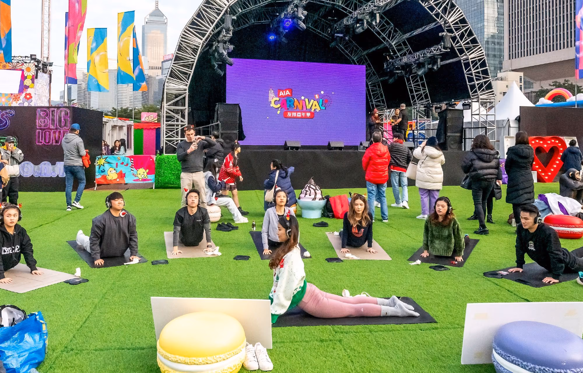 People doing yoga on mats on green grass in front of a stage with a large screen displaying 'AIA Carnival'.