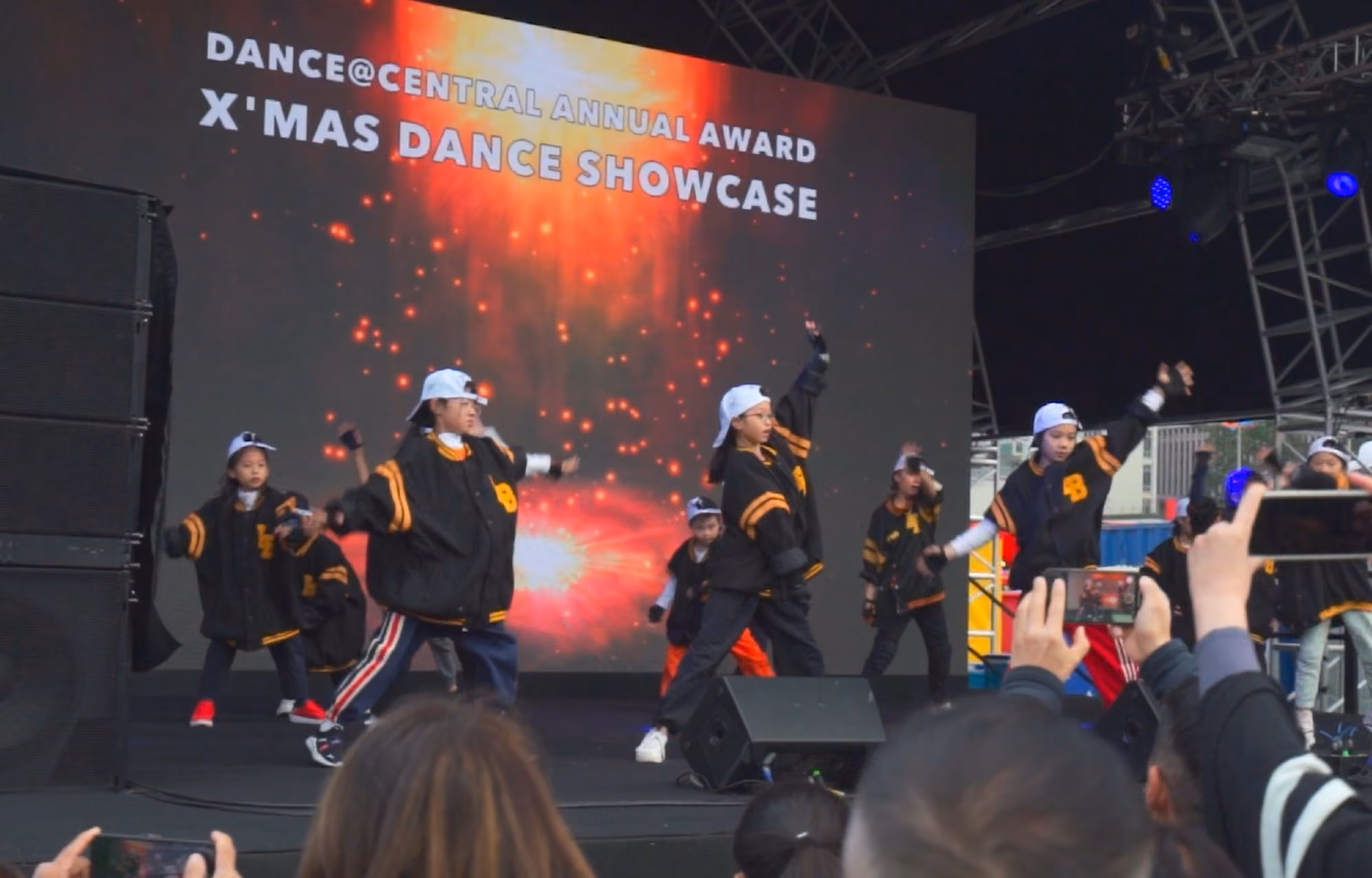 Children in black and orange dance uniforms and white caps performing on stage at the Dance@Central Annual Award X'Mas Dance Showcase with audience filming.