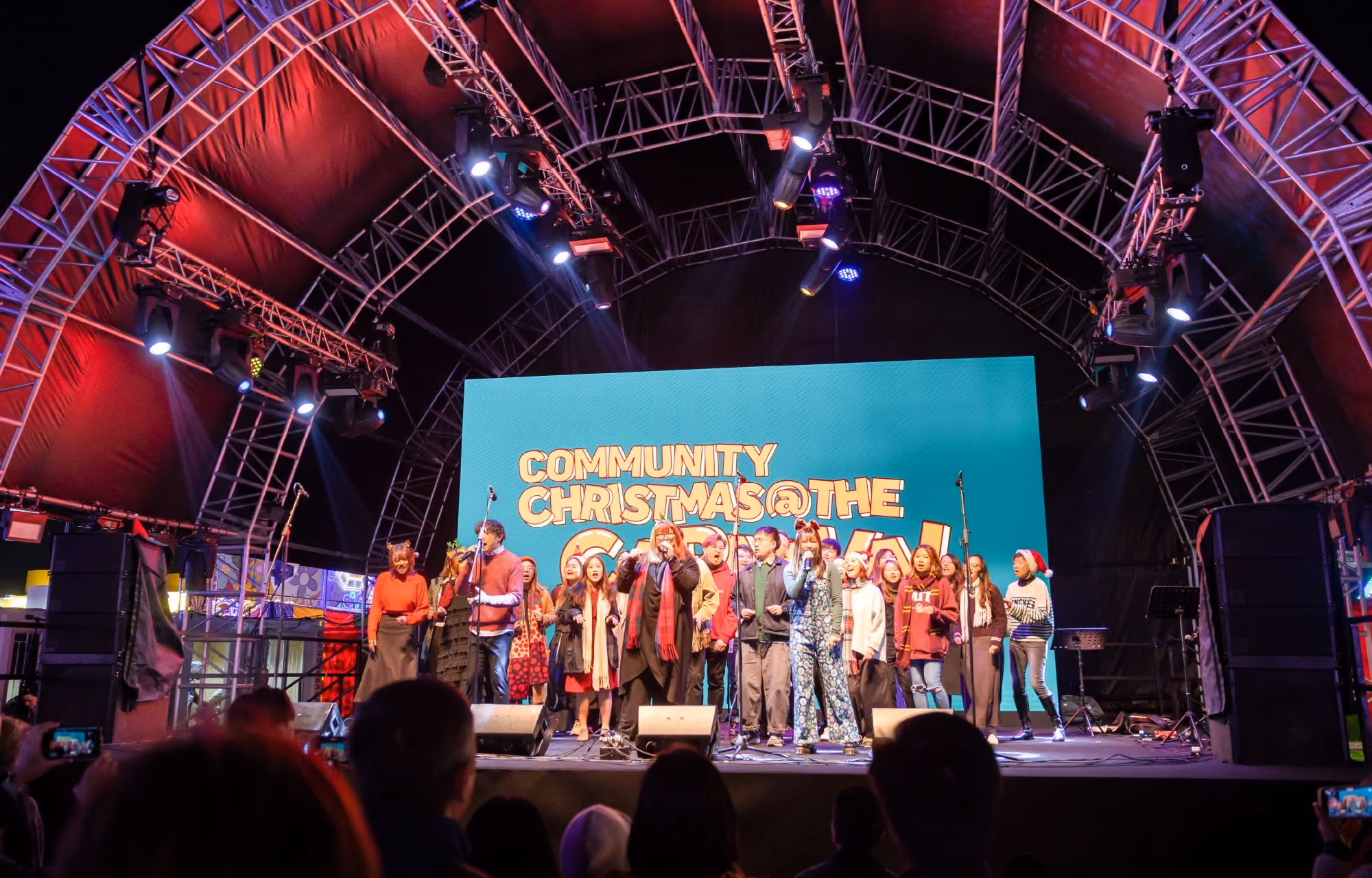 Group of people singing on stage under a metal arch with a screen behind them displaying 'Community Christmas@The'.