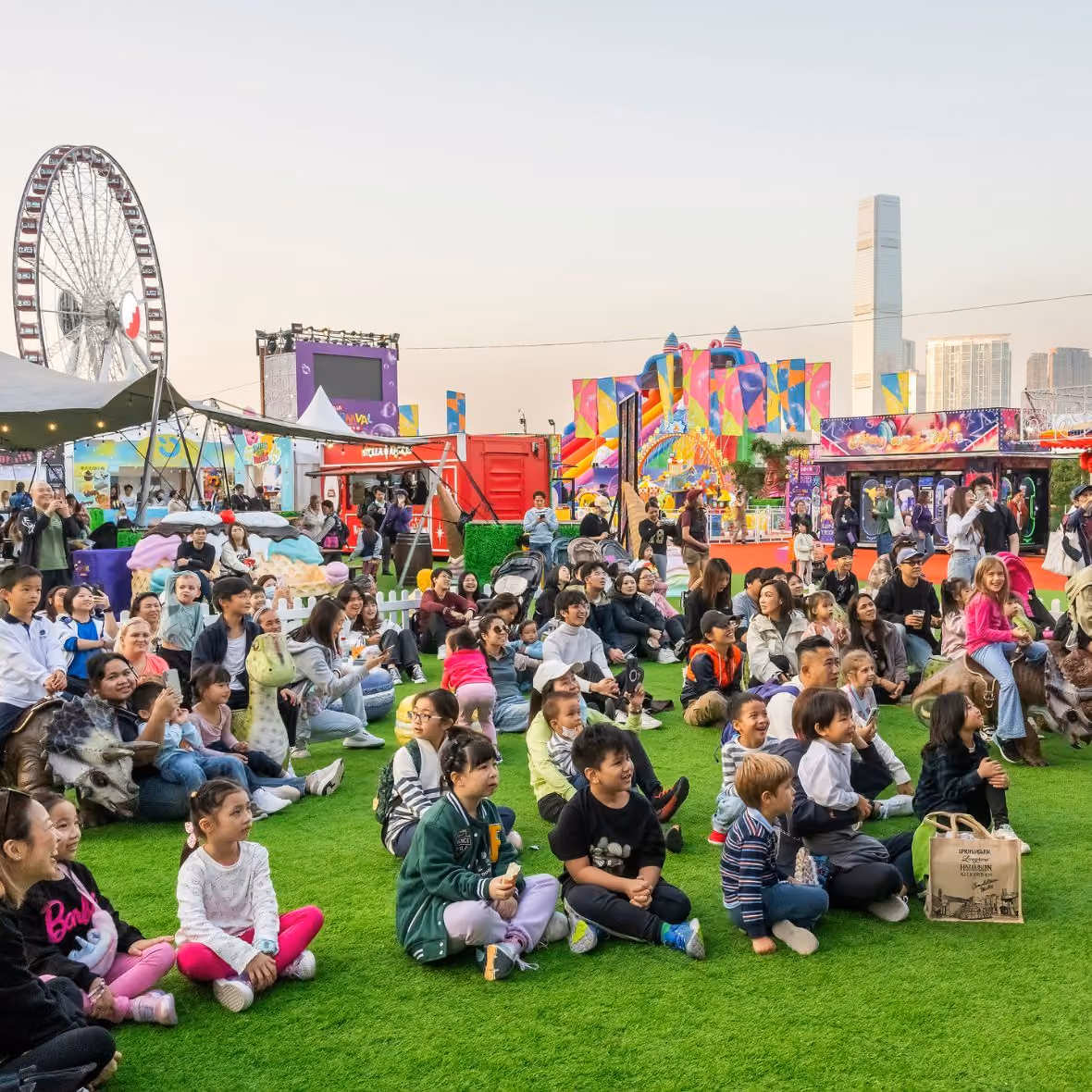 A diverse group of children and adults sitting on green grass at an outdoor event with colorful amusement rides and a Ferris wheel in the background.