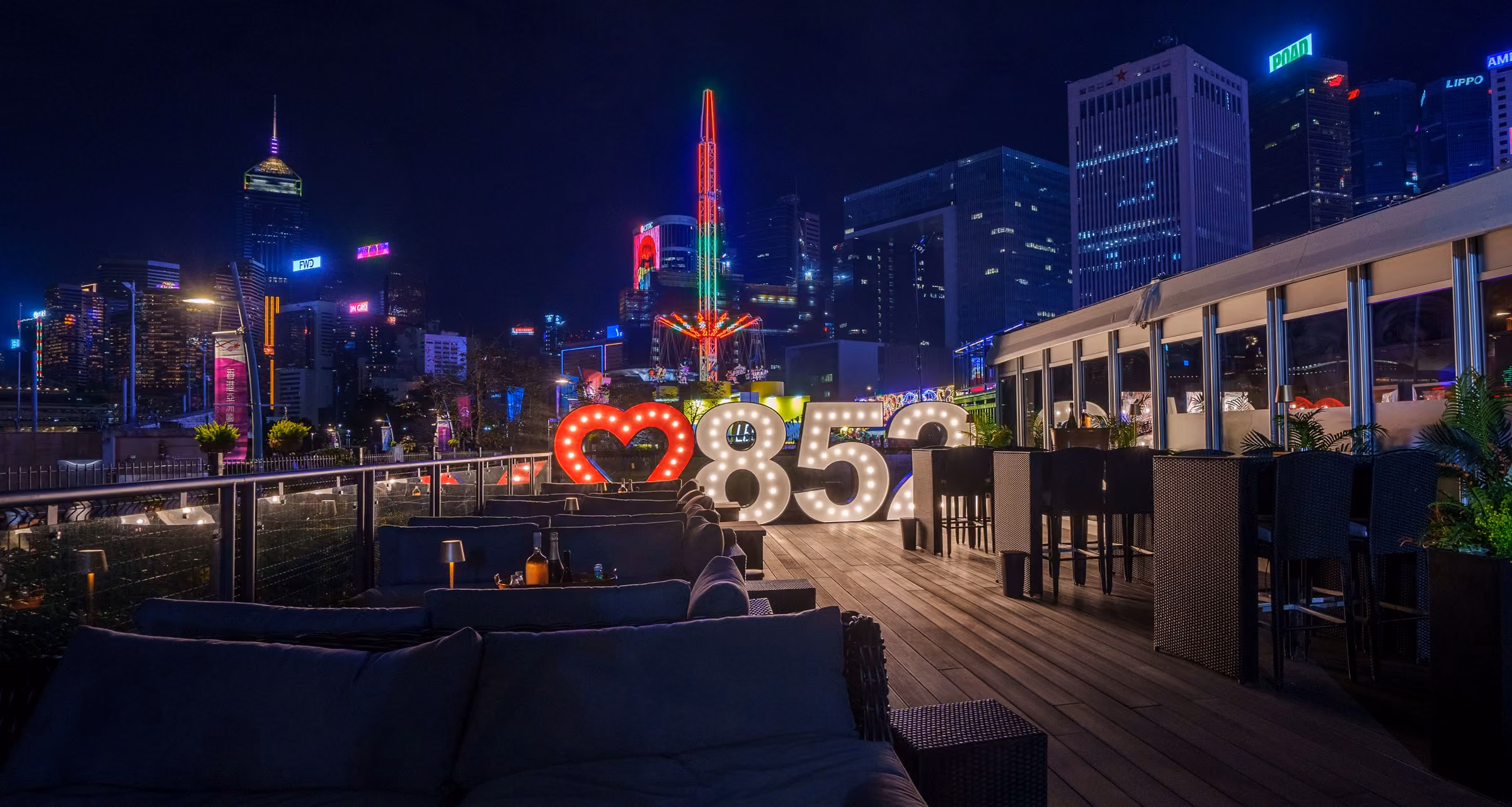 Outdoor rooftop lounge at night with illuminated '852' sign and city skyline in the background.