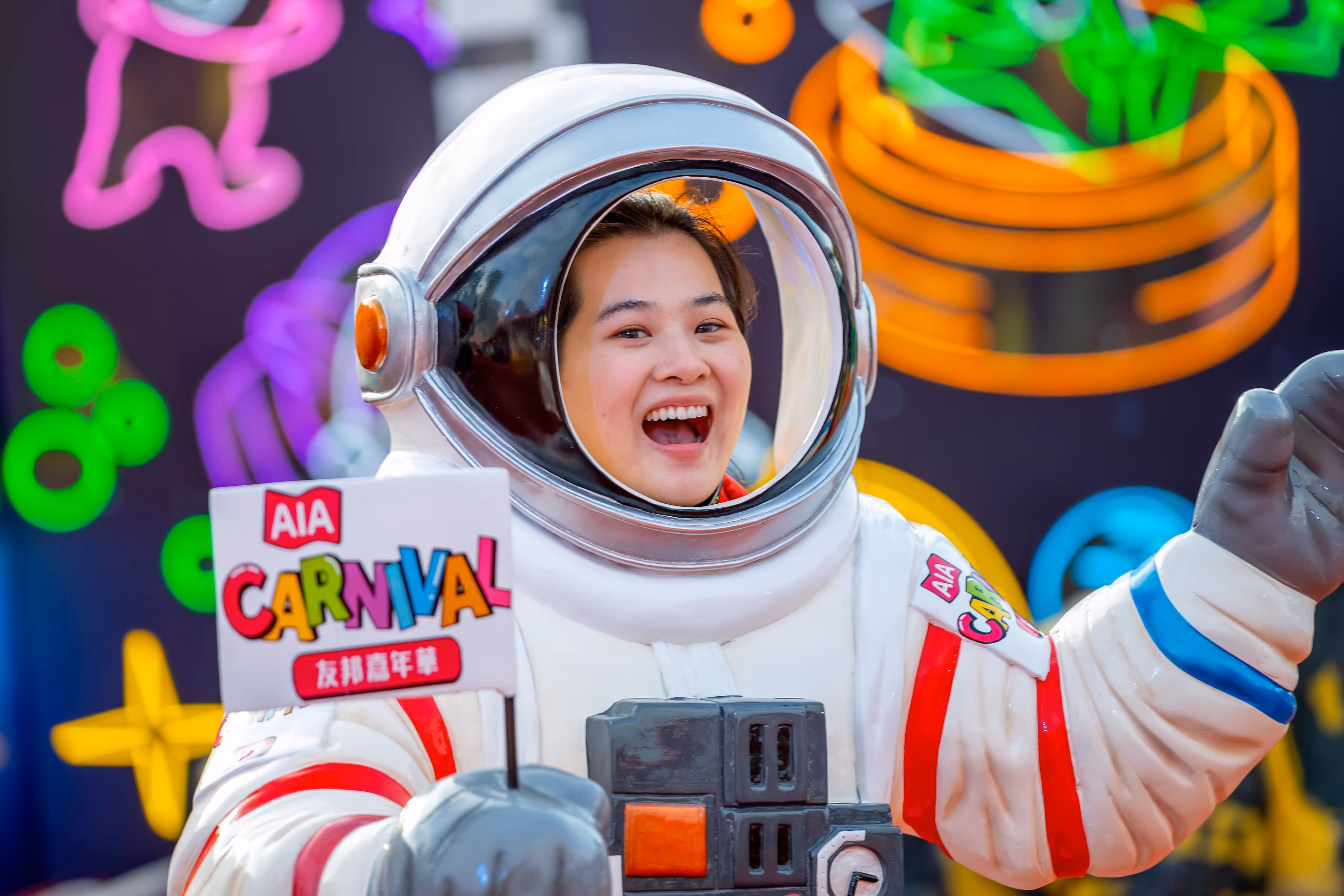 Person smiling inside a white astronaut suit holding a colorful AIA Carnival sign with neon shapes in the background.