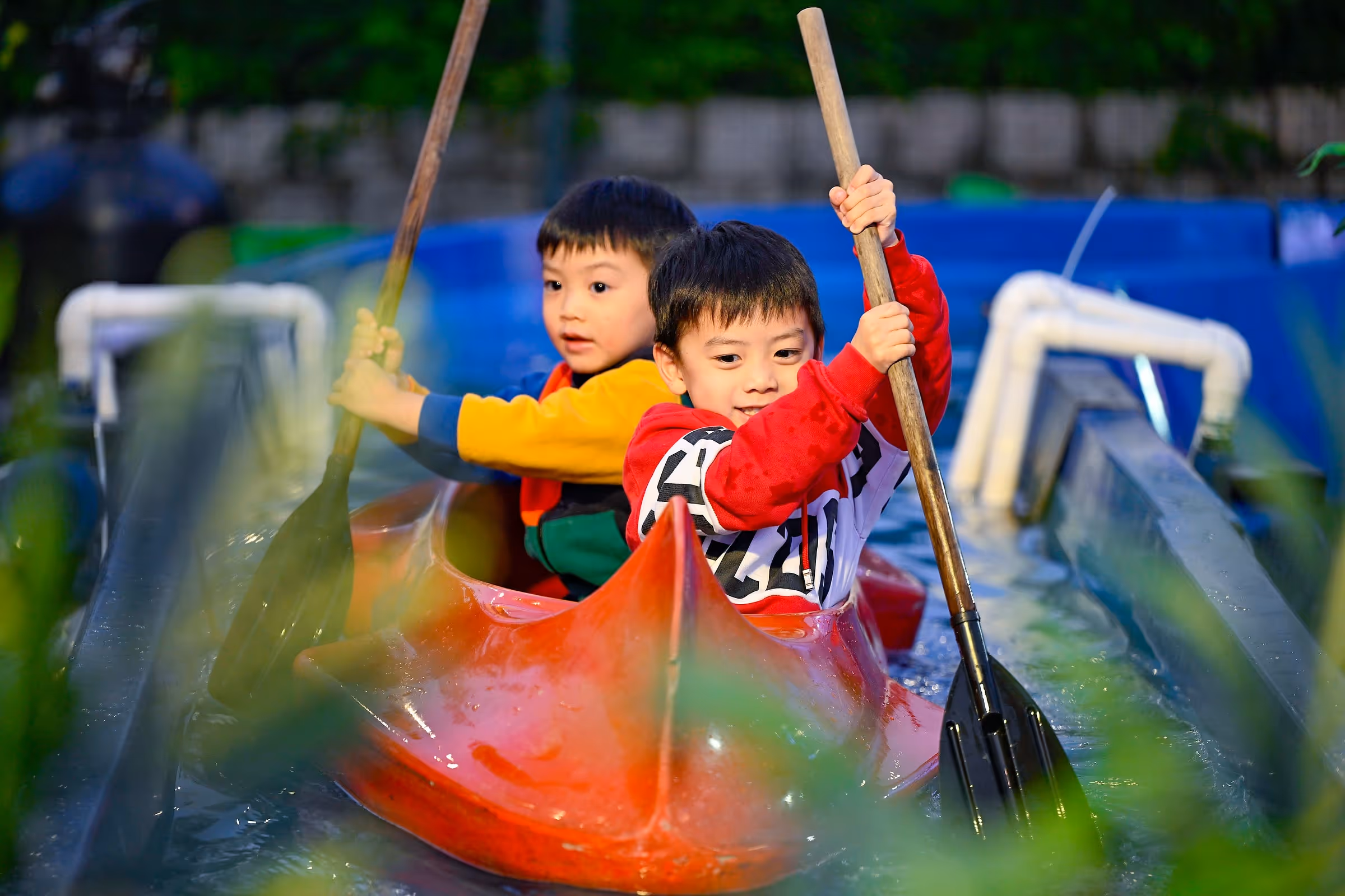 Two young boys paddling together in a red canoe on a narrow water channel.