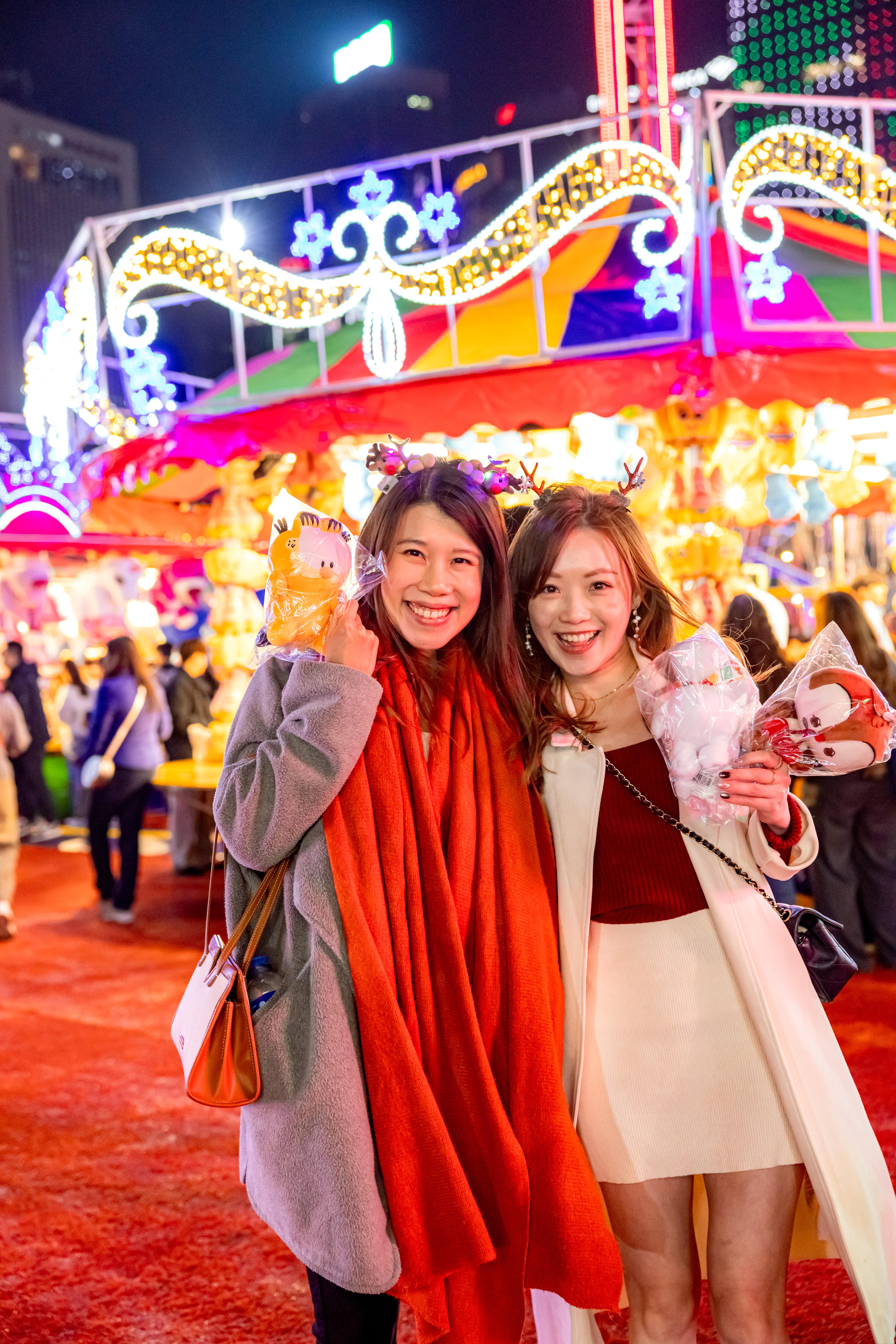 Two women smiling and holding stuffed toys at a brightly lit nighttime carnival or fair.