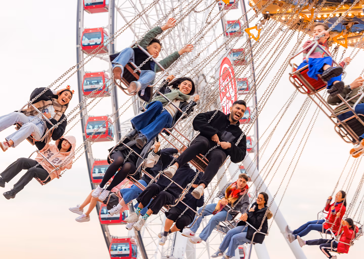 People of various ages enjoying a swing ride at an amusement park with a Ferris wheel in the background.
