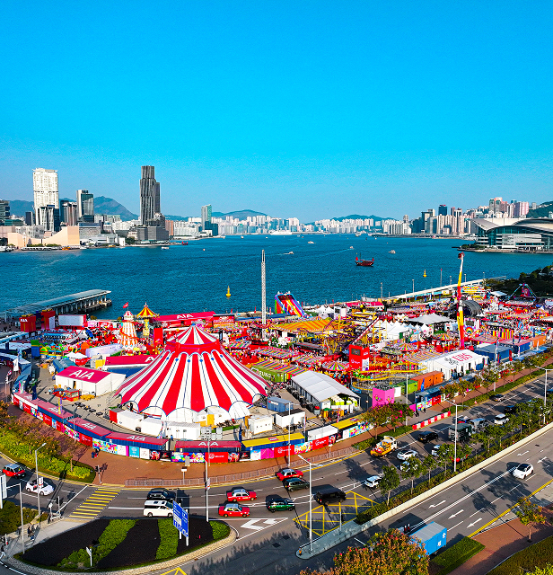 A colorful carnival with red and white tents by a waterfront, set against a skyline of tall buildings under a clear blue sky.