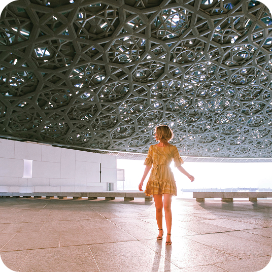 Woman in yellow dress standing under a large geometric patterned ceiling with sunlight in the background.