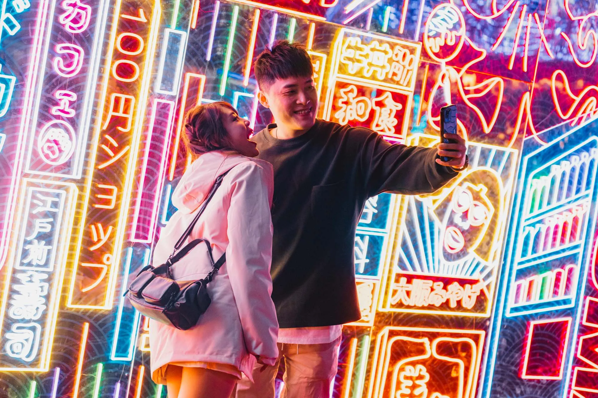 Young couple taking a selfie in front of colorful neon signs with Japanese characters.