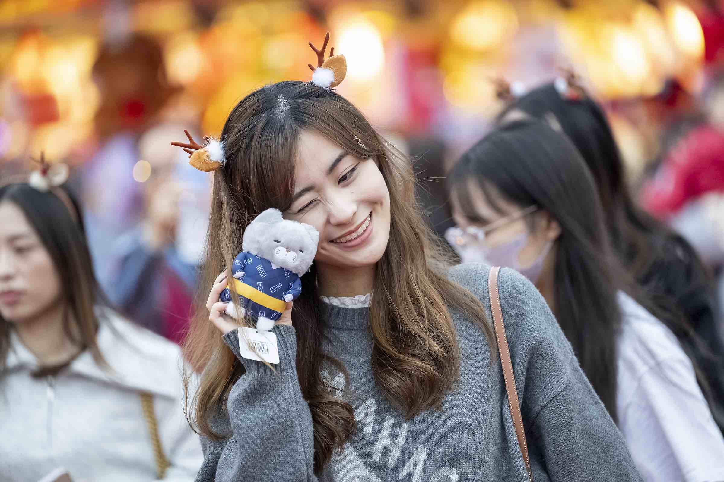 Smiling woman with reindeer antler headband holding a small plush toy next to her face in a crowded outdoor setting.