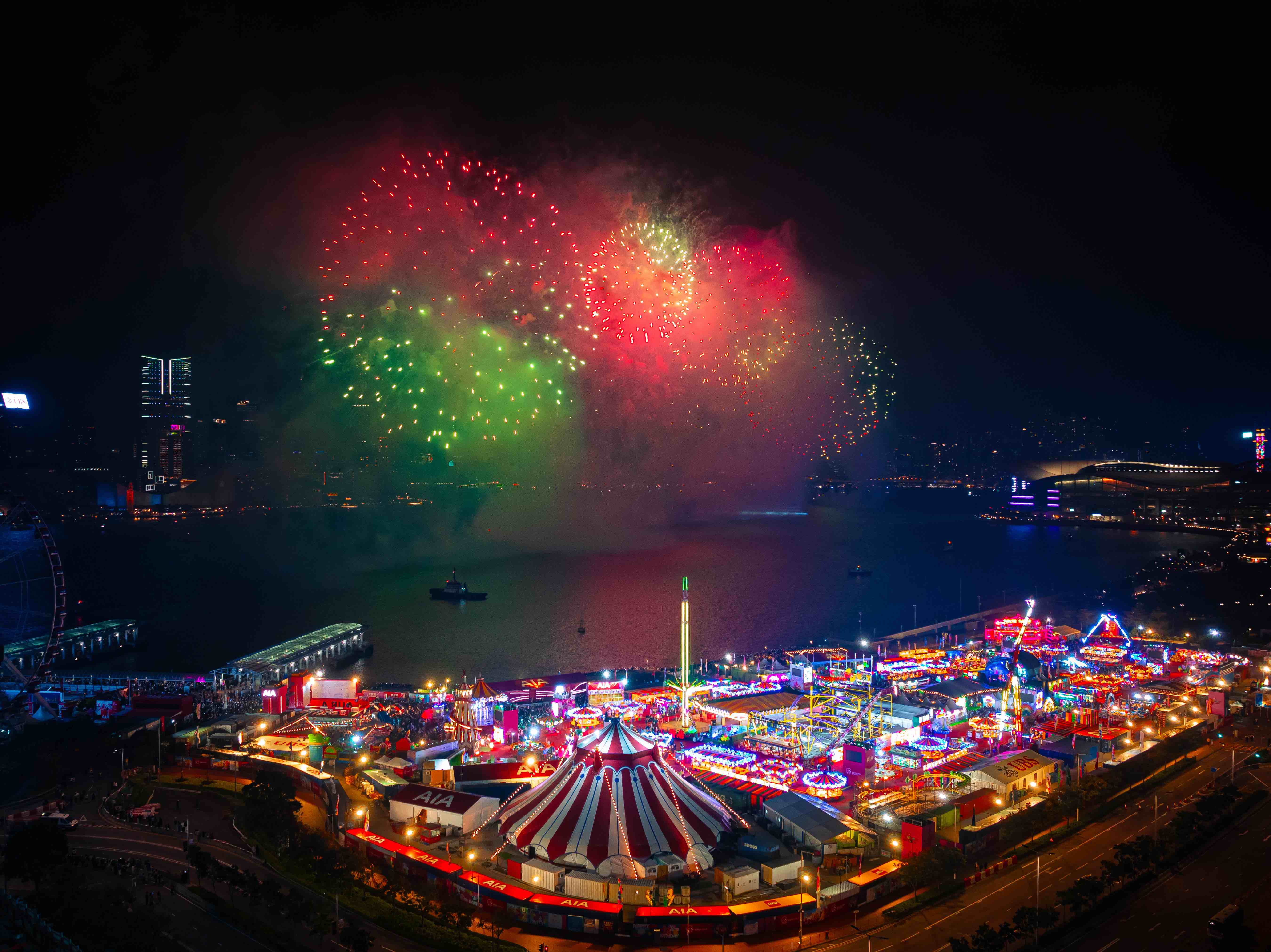 Nighttime carnival by the waterfront with colorful fireworks lighting up the sky above.