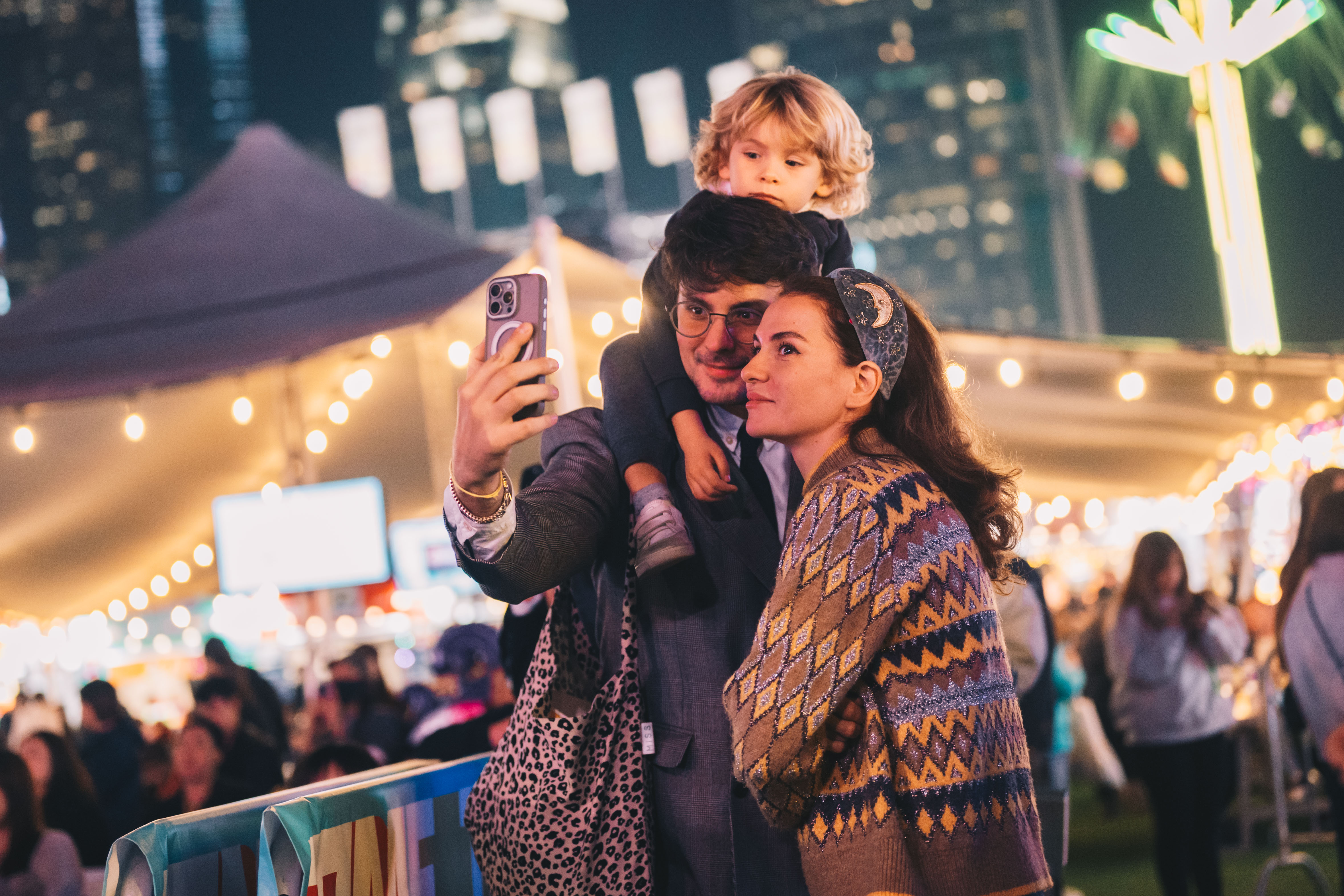 Family of three with child on father's shoulders taking a selfie at a nighttime outdoor event with festive lights.