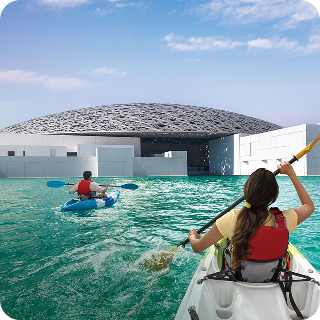 Two people kayaking in turquoise water near a modern building with a patterned dome roof under a partly cloudy sky.