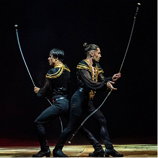 Two male circus performers dressed in black with gold accents holding and spinning long sticks on a dimly lit stage.