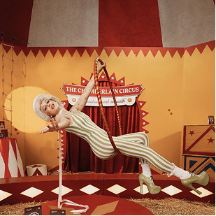 Circus performer in striped jumpsuit and platform heels balancing on a pole inside a vintage circus tent.