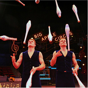 Two male jugglers in black sleeveless vests juggling clubs under a Big Apple Circus sign.