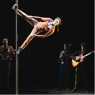 Performer in pink outfit doing a horizontal pole hold with three musicians playing instruments in the background on a dark stage.