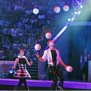 Two circus performers juggling multiple balls on stage in front of an audience.