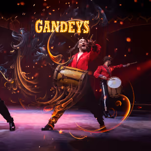 Two performers in red shirts playing traditional drums with energetic poses on stage under a lit sign that says 'GANDEYS'.