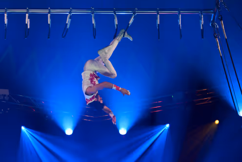 Aerial performer dressed in white and red hanging upside down from metal bars with blue stage lights in the background.
