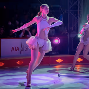 Figure skater in a white costume performing on ice under colorful lights with an audience in the background.