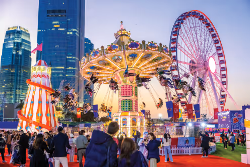 Evening scene of a lively carnival with a brightly lit swing ride, a large Ferris wheel, a tall striped slide, and a crowd of people against a city skyline.