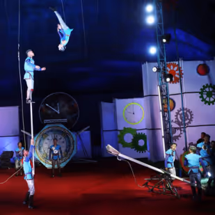 Circus performers executing a trampoline acrobatic stunt under a blue tent with gears and clock decorations in the background.