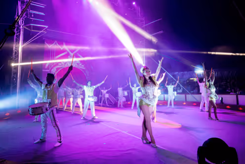 Performers in white costumes dancing and playing drums on a stage with purple lighting and spotlights.