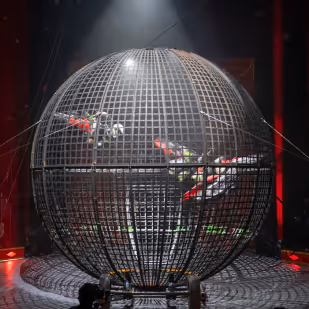 Two motorcyclists performing stunts inside a large spherical metal cage under spotlight.