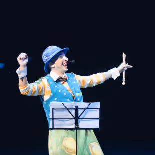 Clown in colorful costume and hat enthusiastically performing on stage with a music stand in front.