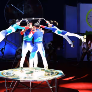 Four performers in colorful outfits roller skate on a rotating platform, two are airborne holding sticks while the other two support them.