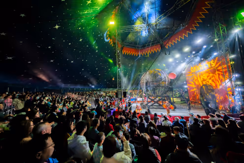 A large indoor circus audience watches a brightly lit stage with performers and a spherical metal cage under a star-patterned tent ceiling.