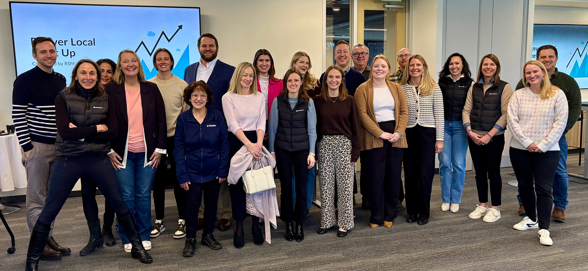 Group of 20 professionals standing and smiling in an office with a presentation screen displaying a rising graph in the background.