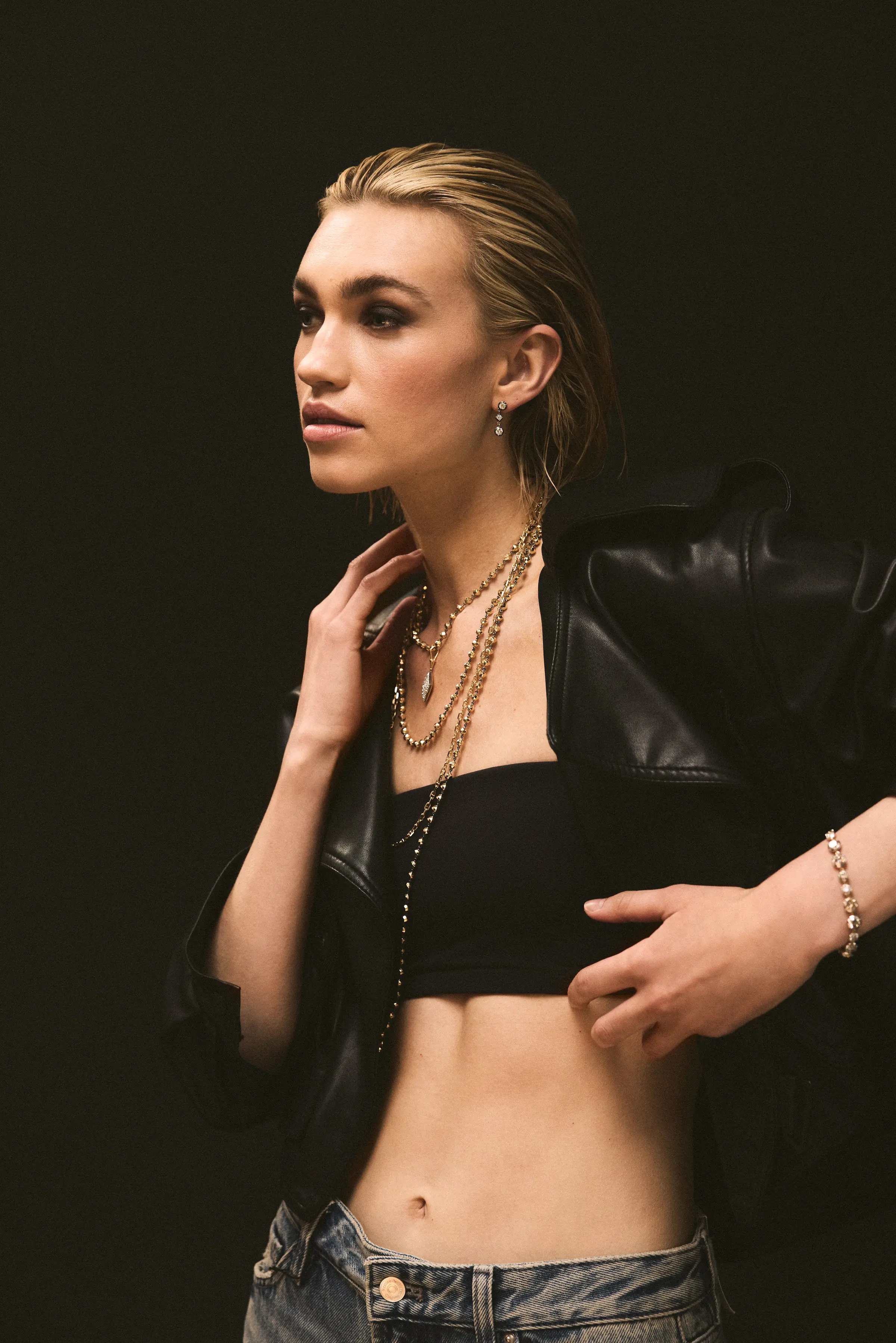 Fashion model with slicked-back hair wearing layered gold necklaces, earrings, a black cropped top, a black leather jacket, and blue jeans against a dark background.