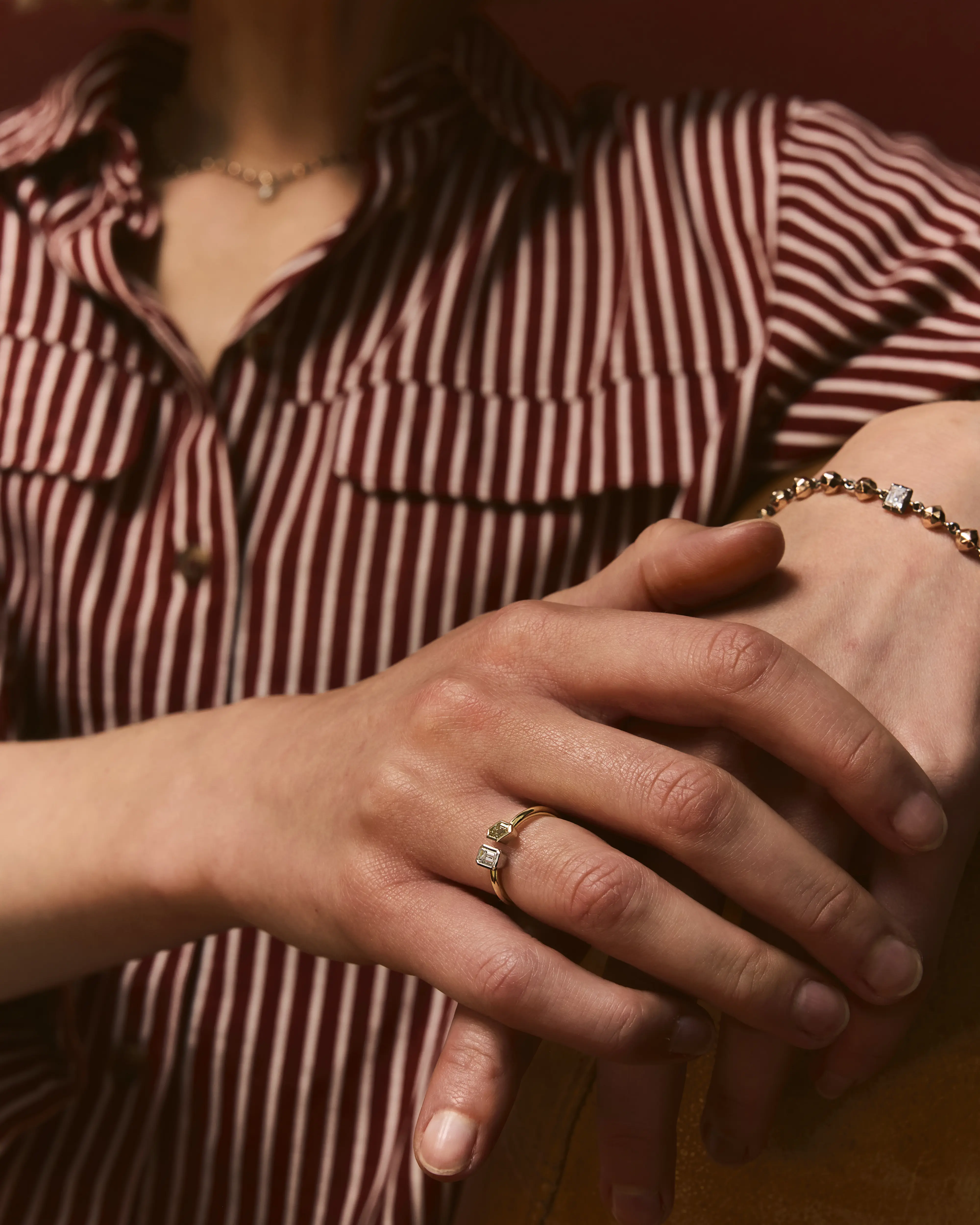 Close-up of a person wearing a red and white striped shirt showing their hands adorned with a gold ring with two gemstones and a gold bracelet.