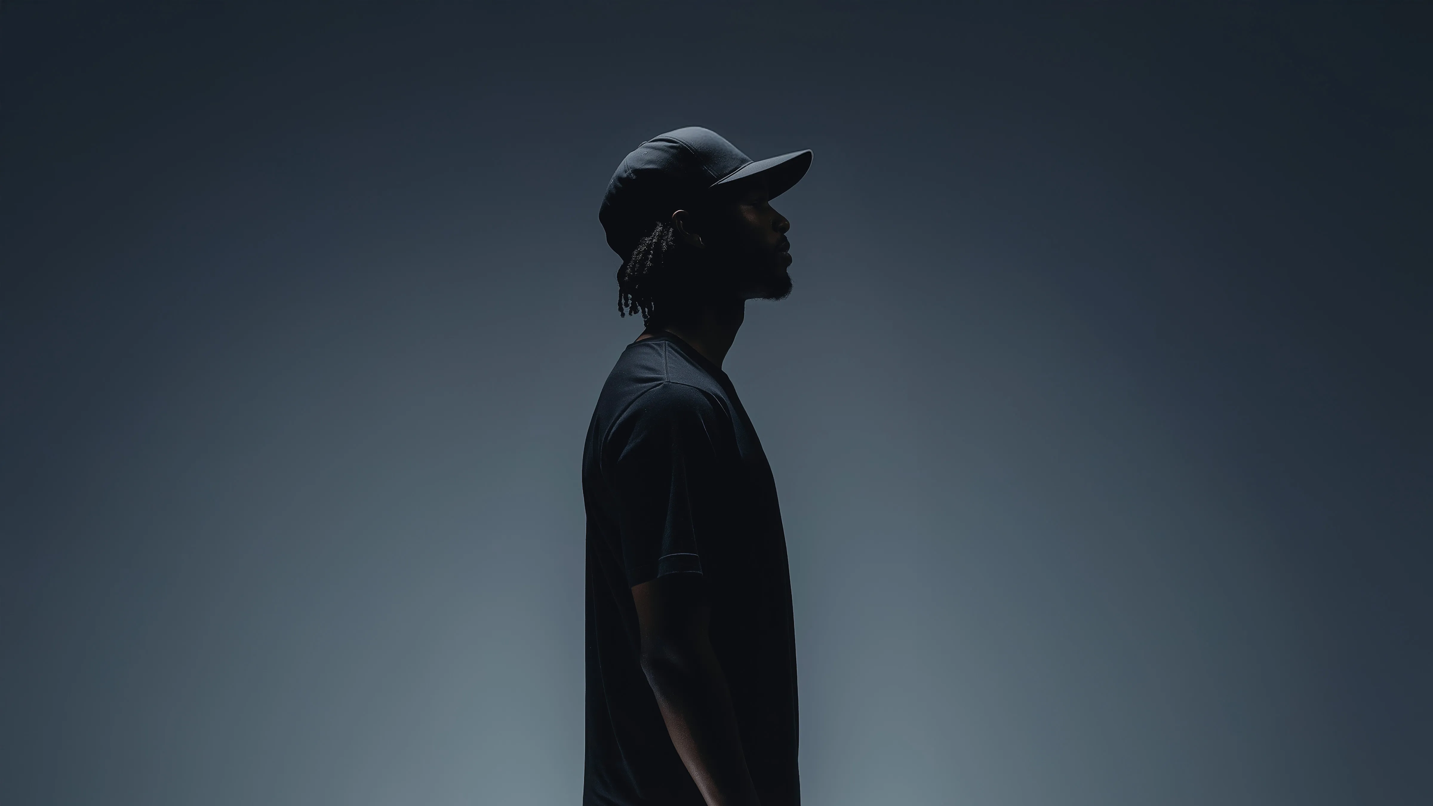 Side profile silhouette of a man wearing a black cap and black shirt against a dark gradient background.