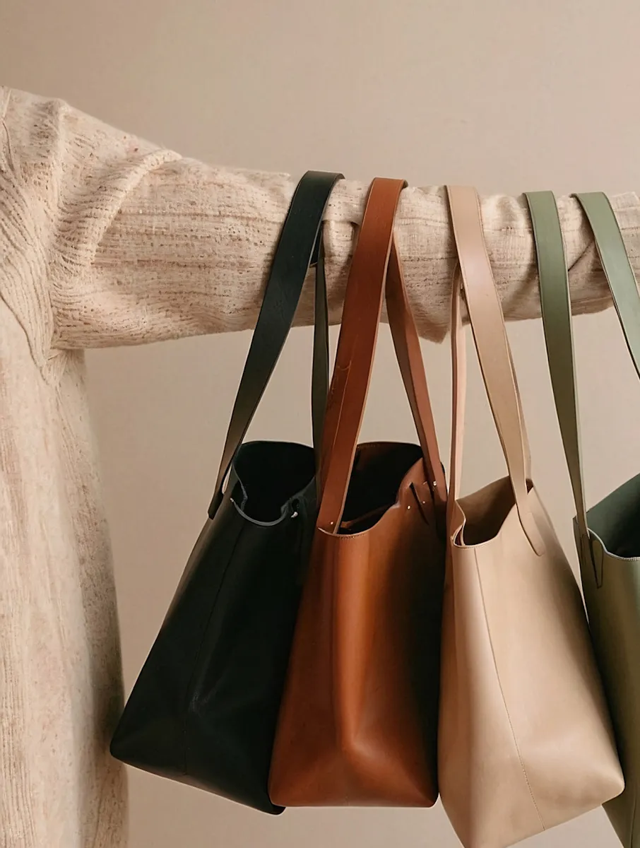 Arm in a beige sweater holding four leather tote bags in black, brown, beige, and olive green.