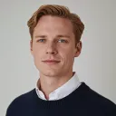 Portrait of a young man with blond hair wearing a white collared shirt and dark sweater against a plain gray background.