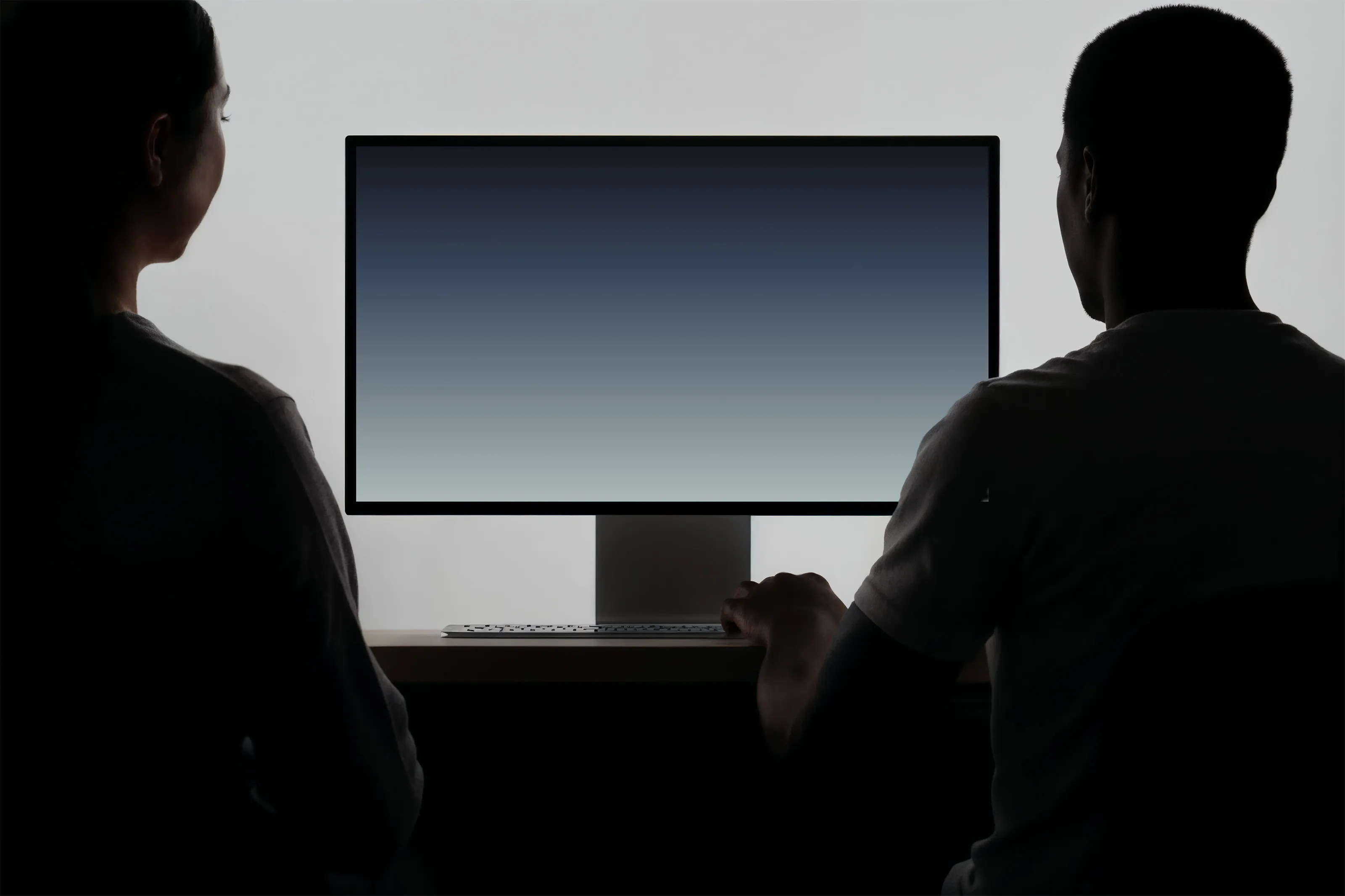 Silhouettes of two people sitting side by side, looking at a large computer monitor with a gradient blue screen.
