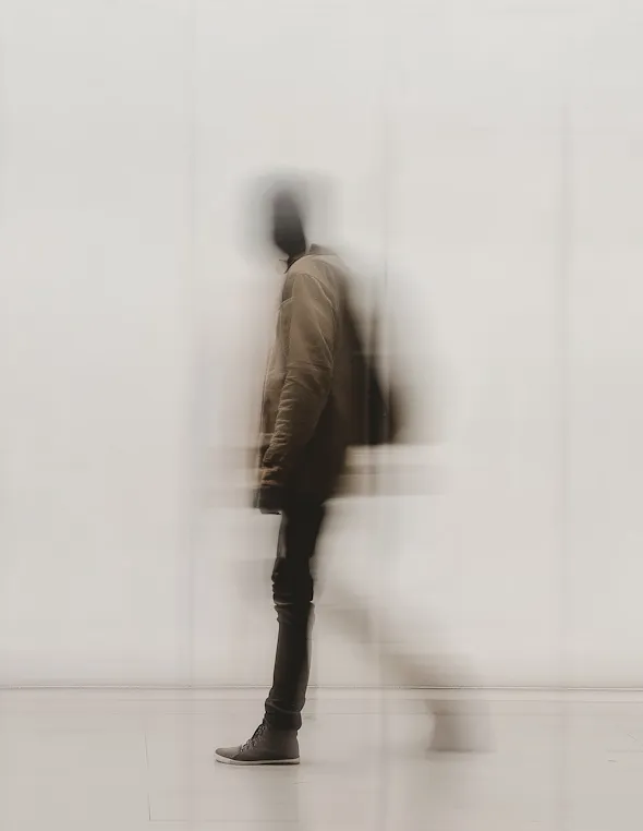 Blurred image of a person wearing a brown jacket and black pants standing against a plain light background.