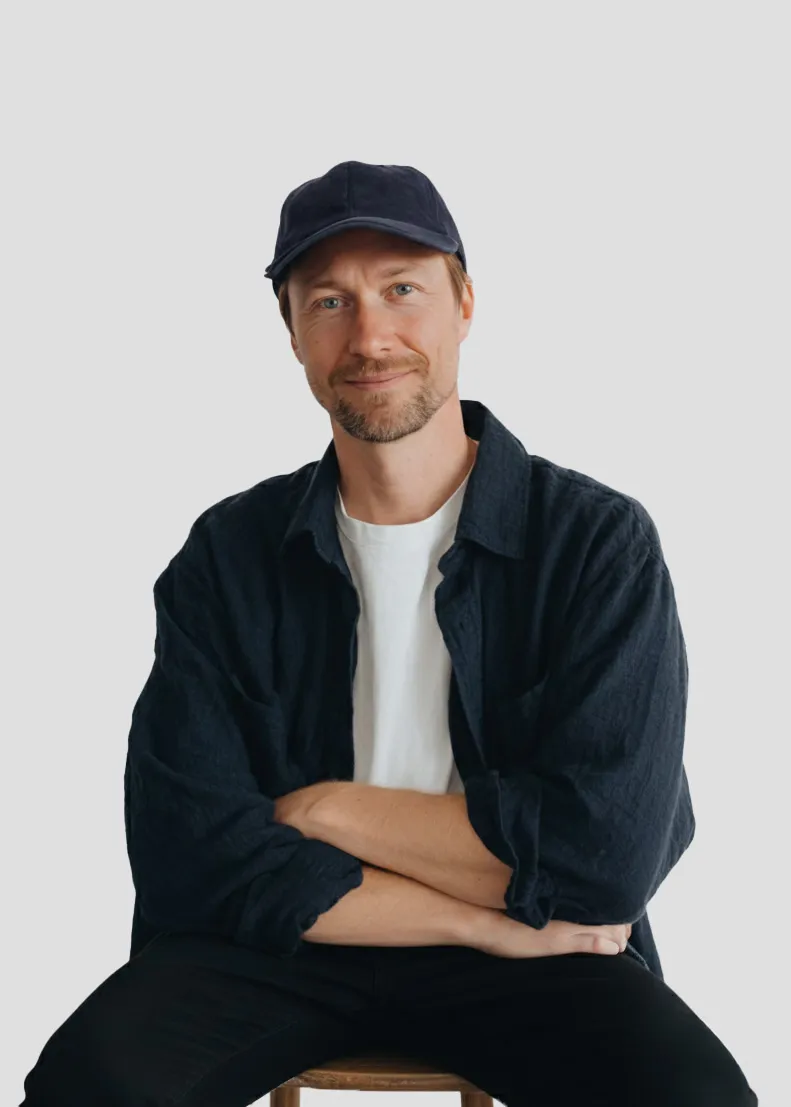 Man with light beard wearing a black cap, black shirt over a white t-shirt, sitting with arms crossed against a plain light background.