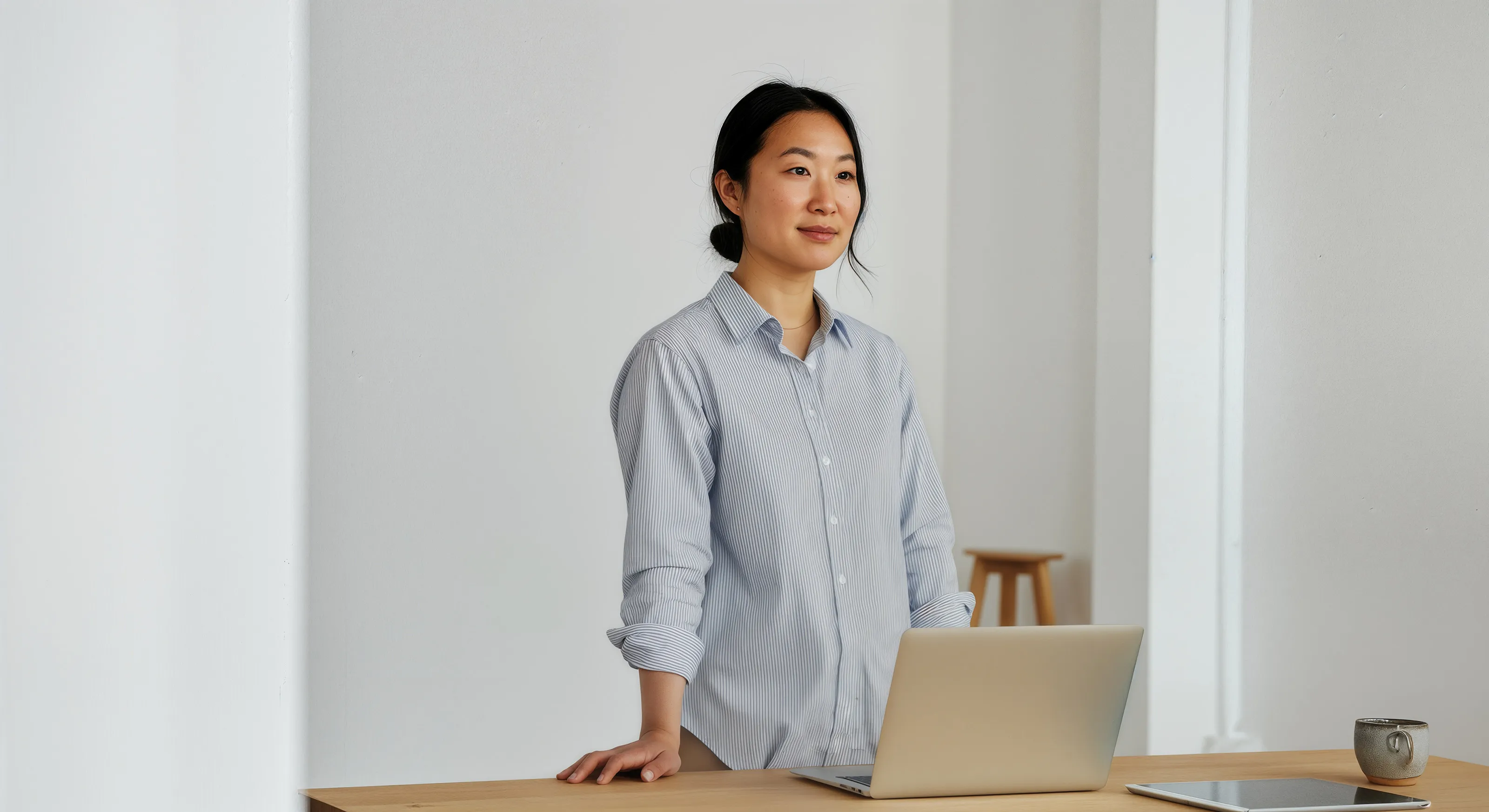 Young woman in a striped shirt standing at a desk with a laptop, tablet, and coffee cup in a minimalist room.